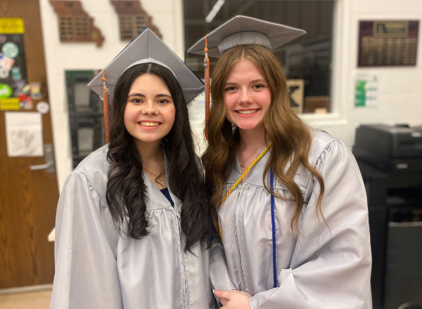  Two graduates smile while looking at the camera in gray graduation caps and gowns